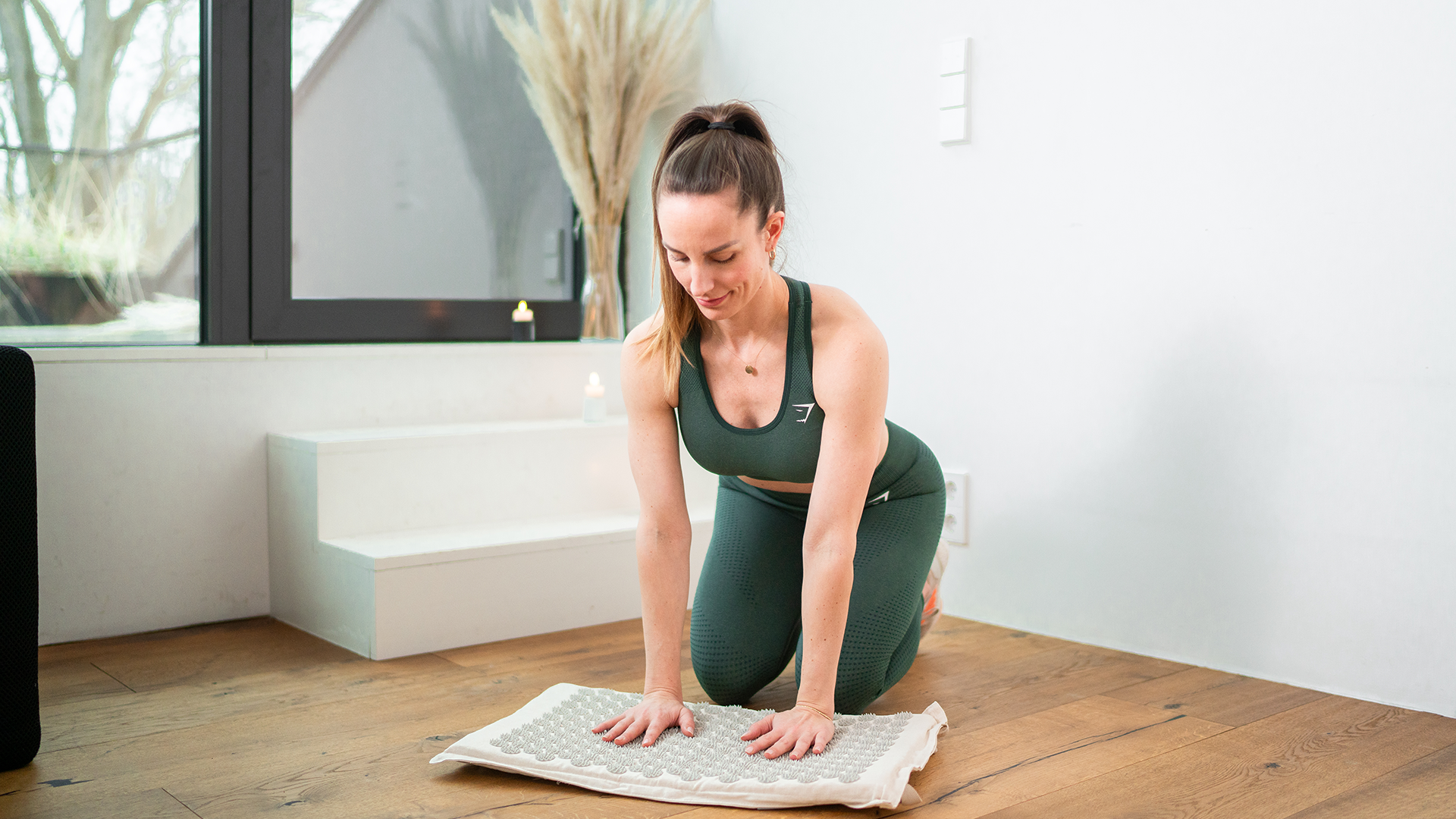 Eine Frau in grüner Sportkleidung kniet auf dem Boden und legt ihre Hände auf eine Akupressurmatte. Sie bereitet sich auf eine Entspannungs- oder Massageübung vor. Im Hintergrund sind Kerzen und eine minimalistische Einrichtung zu sehen, was eine ruhige und entspannte Atmosphäre schafft. Die Szene zeigt, wie die Matte zur Verbesserung des Wohlbefindens genutzt wird.