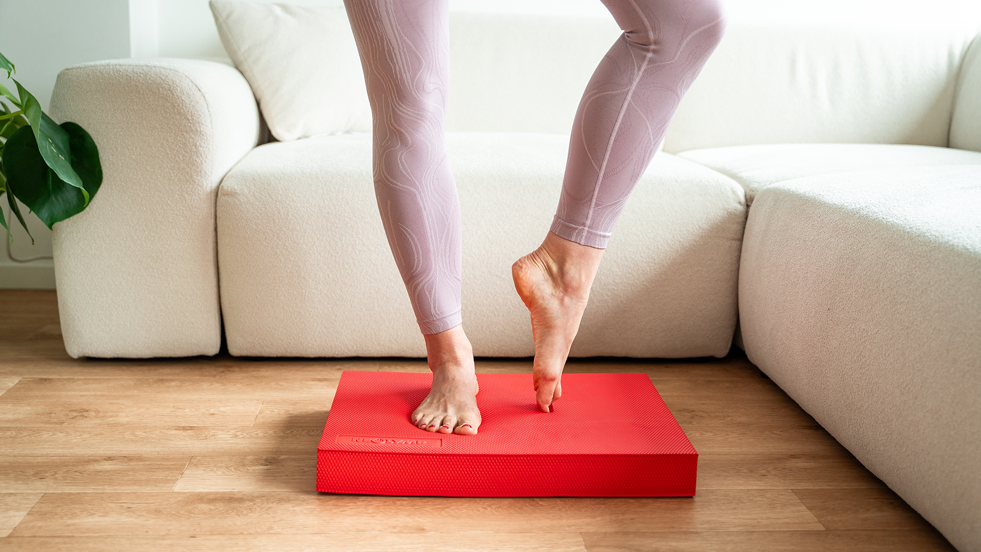 Eine Person steht auf Zehenspitzen auf einer roten Neolymp-Matte, die auf einem Holzboden vor einem cremefarbenen Sofa liegt. Die Person trägt eine lilafarbene, eng anliegende Sporthose, und der Fokus liegt auf den Füßen und Unterschenkeln.