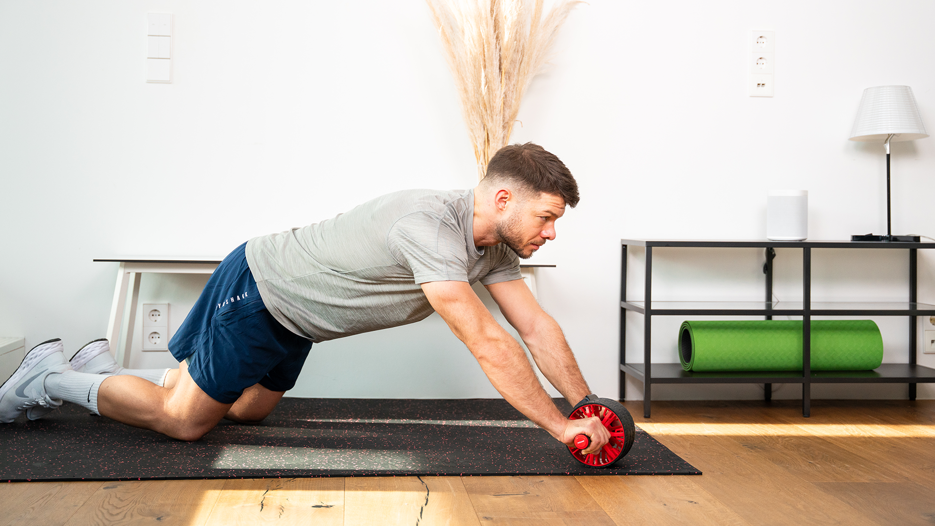 Mann trainiert mit einem Bauchroller auf einer schwarzen Fitnessmatte in kniender Position, trägt graues T-Shirt und blaue Shorts in einem modern eingerichteten Fitnessraum.