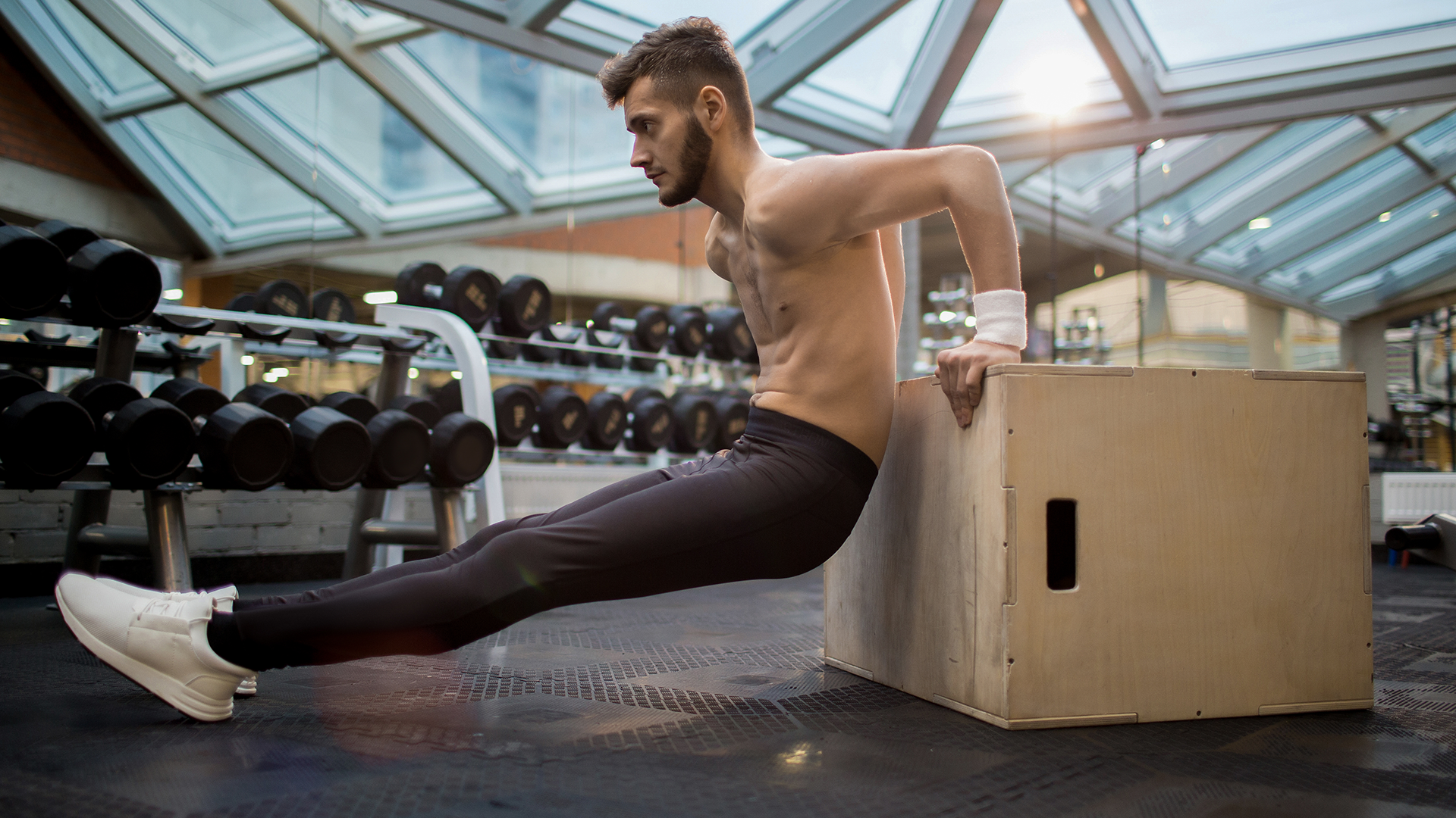 Mann in schwarzer Sportkleidung macht Trizeps-Dips auf einer Holzbox im Fitnessstudio. Im Hintergrund sind Hanteln und große Fenster zu sehen.