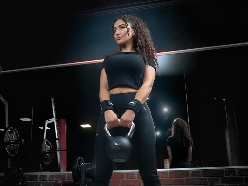 Junge Frau im Fitnessstudio hält eine Kettlebell in den Händen und schaut selbstbewusst in die Ferne, bereit für ihr Krafttraining.