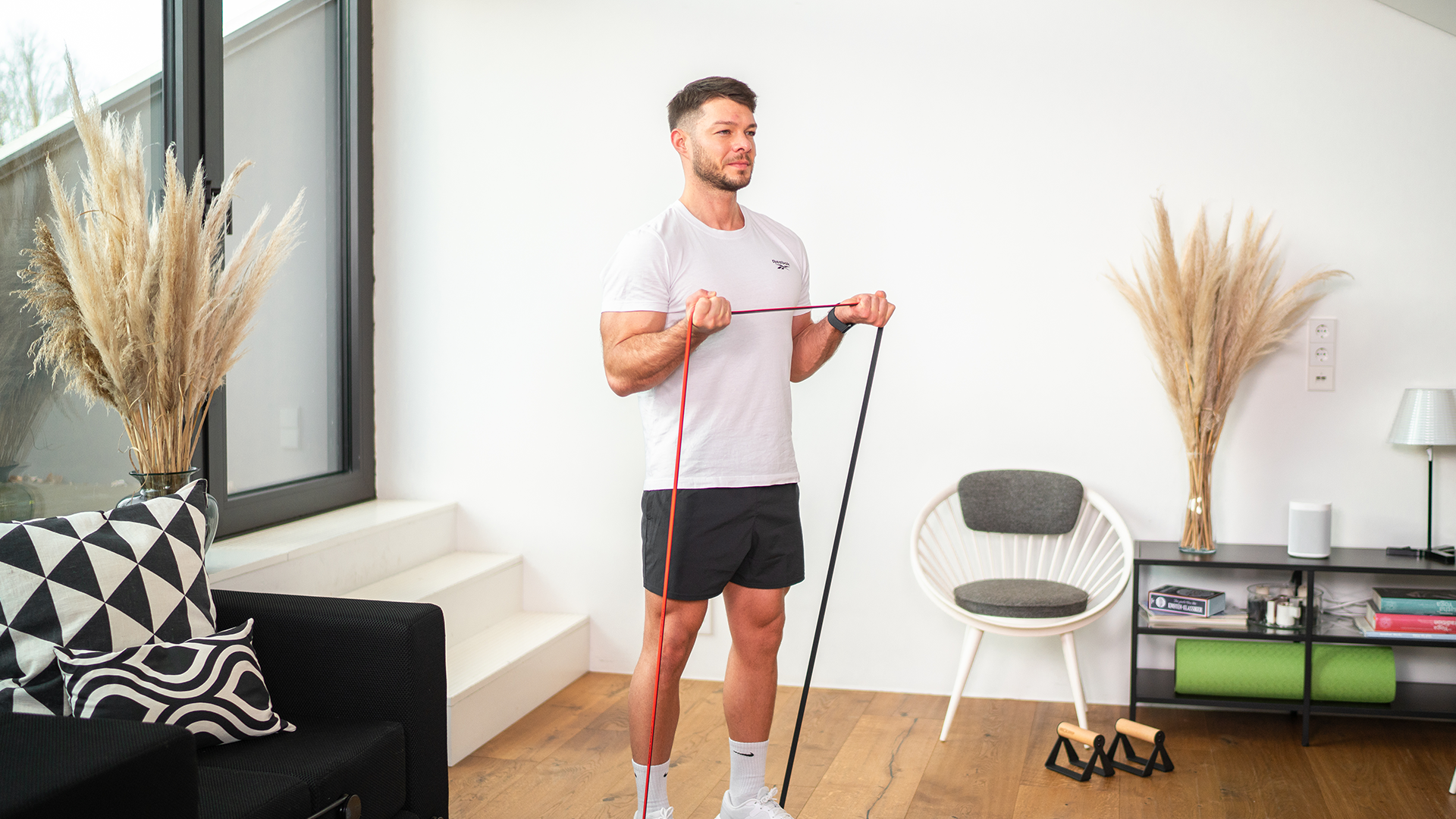 Mann führt Bizeps-Curls mit einem Neolymp Widerstandsband in einem modernen, minimalistisch eingerichteten Wohnzimmer durch, während er ein weißes T-Shirt und schwarze Shorts trägt.