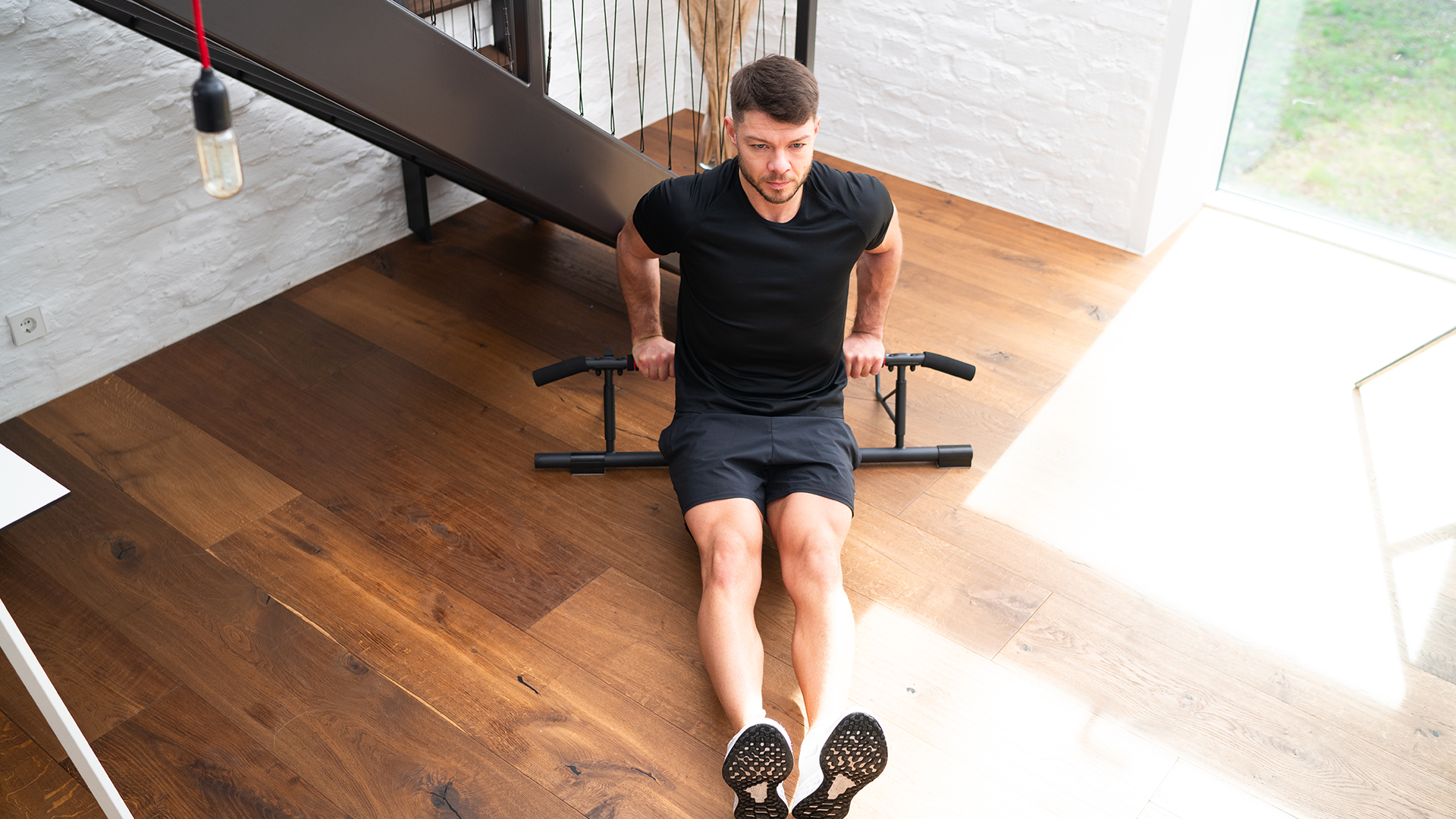 Mann führt Dips auf einem Fitnessgerät in einem modernen Raum mit Holzfußboden aus, trägt ein schwarzes T-Shirt und Shorts, und verwendet die Dips-Stange für ein Trizeps-Workout.