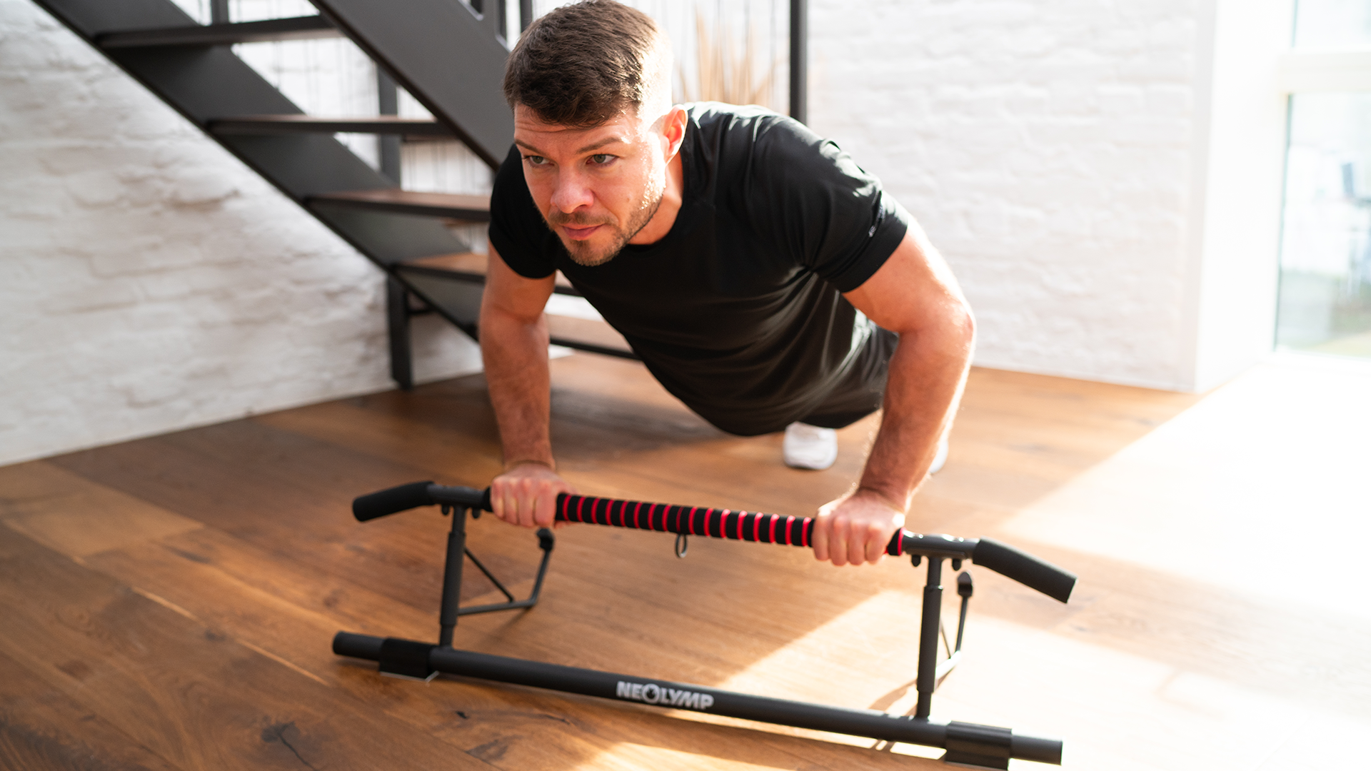 Mann führt Liegestütze auf einem Neolymp Push-up-Barren aus, während er auf einem Holzboden in einem modernen Fitnessraum trainiert und dabei ein schwarzes T-Shirt trägt.