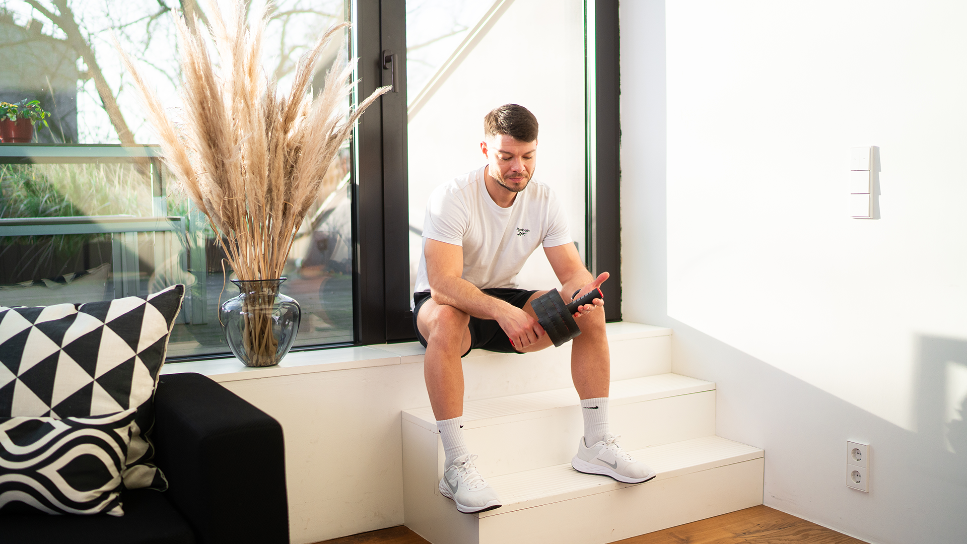 Ein Mann sitzt entspannt auf einer Treppe in einem modernen Raum mit großen Fenstern und natürlichem Licht. Er hält einen Ab-Roller in den Händen und scheint eine Pause von seinem Training zu machen. Der Raum ist stilvoll eingerichtet, mit einer Vase voller Pampasgras und dekorativen Kissen, was eine angenehme und ruhige Atmosphäre vermittelt. Die Szene zeigt einen Moment der Entspannung und Selbstreflexion nach einem Workout.