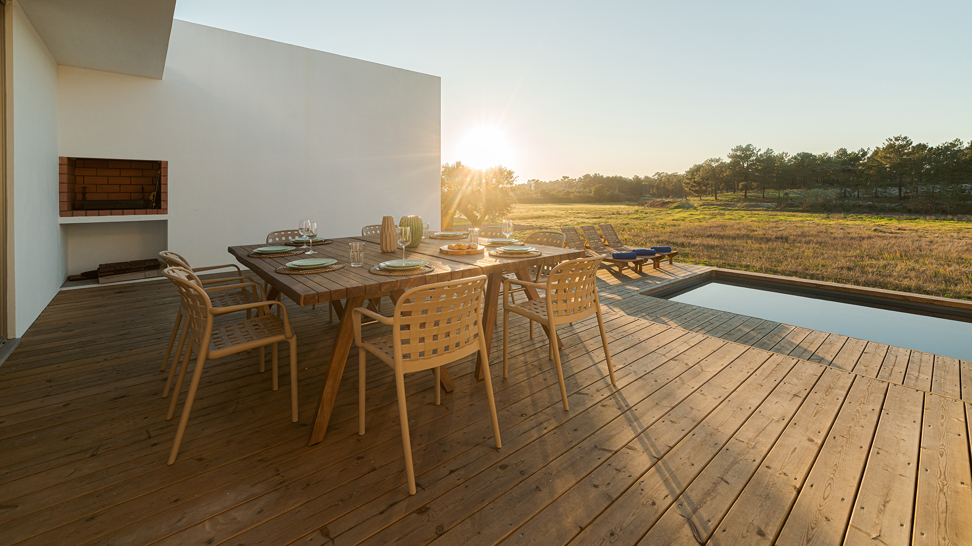 Moderne Terrasse mit einem großen Holztisch, umgeben von beige geflochtenen Stühlen, gedeckt für sechs Personen. Im Hintergrund sind Liegestühle neben einem rechteckigen Pool zu sehen. Die Sonne geht über einer weiten Wiesenlandschaft unter, während die Szenerie in ein warmes, goldenes Licht getaucht ist. Links an der weißen Wand befindet sich ein eingebauter Grill.