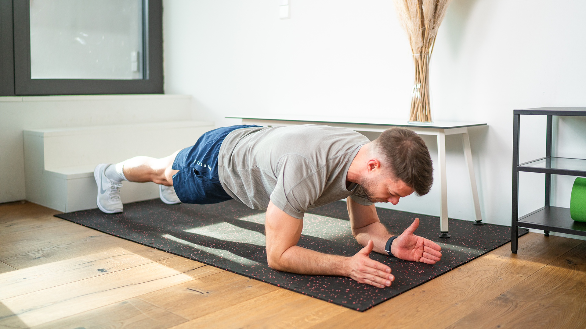 Ein Mann führt eine Unterarmplanke auf einer schwarzen Trainingsmatte mit roten Sprenkeln durch. Er hält die Spannung in einer stabilen Position, während er sich auf seine Körpermitte konzentriert. Die Übung wird in einem hellen, modernen Raum mit Holzfußboden und minimalistischem Interieur ausgeführt, was ein effektives Heimtraining zeigt.