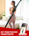 Mit praktischer Tragetasche. Eine Frau trägt einen Gymstick in einer schwarzen Tragetasche von Neolymp über der Schulter. Sie steht in einem hellen Wohnzimmer und trägt rosafarbene Sportkleidung. Im Bild ist auch ein Smartphone mit dem Text "Inklusive Übungen" zu sehen.