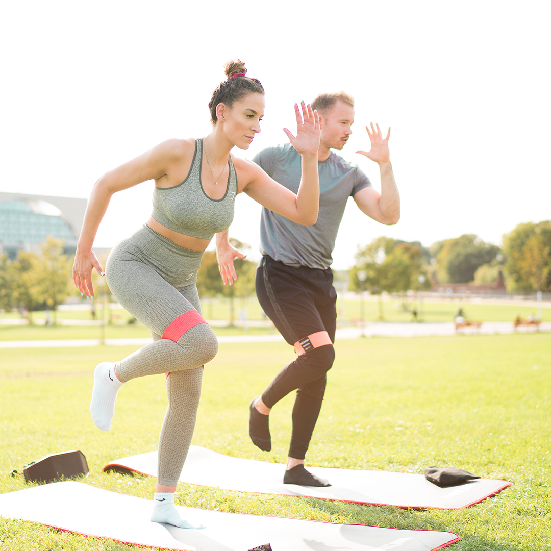 Ein Mann und eine Frau trainieren im Freien auf einer Wiese, beide verwenden rote Widerstandsbänder um ihre Oberschenkel. Sie führen eine Balance-Übung auf Gymnastikmatten aus, während sie konzentriert ihre Arme anheben. Die Szene zeigt eine gemeinsame Fitness-Session im Freien bei sonnigem Wetter, ideal für Kraft- und Ausdauertraining.