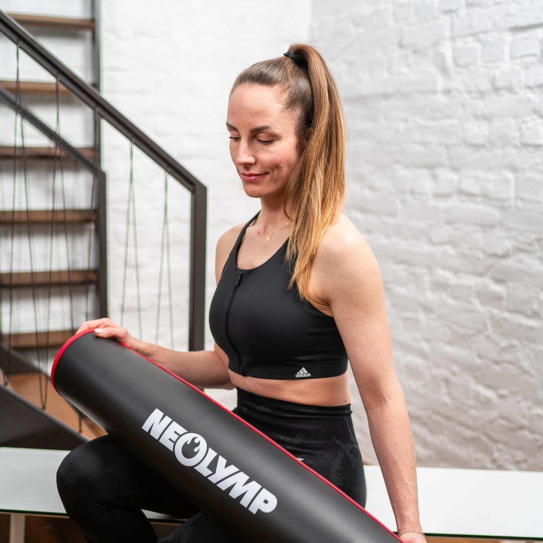 Frau in Sportkleidung hält eine aufgerollte Neolymp Fitnessmatte mit rotem Rand, stehend vor einer Treppe.