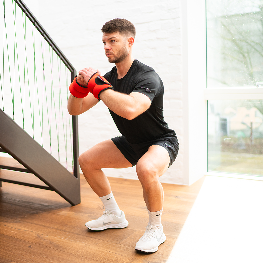 Ein Mann in schwarzer Sportkleidung und weißen Sportschuhen macht Kniebeugen auf einer Treppe. Er trägt orangefarbene Neolymp-Gewichtsbandagen an den Handgelenken, um das Training zu intensivieren.