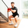 Ein Mann in schwarzer Sportkleidung und weißen Sportschuhen macht Kniebeugen auf einer Treppe. Er trägt orangefarbene Neolymp-Gewichtsbandagen an den Handgelenken, um das Training zu intensivieren.