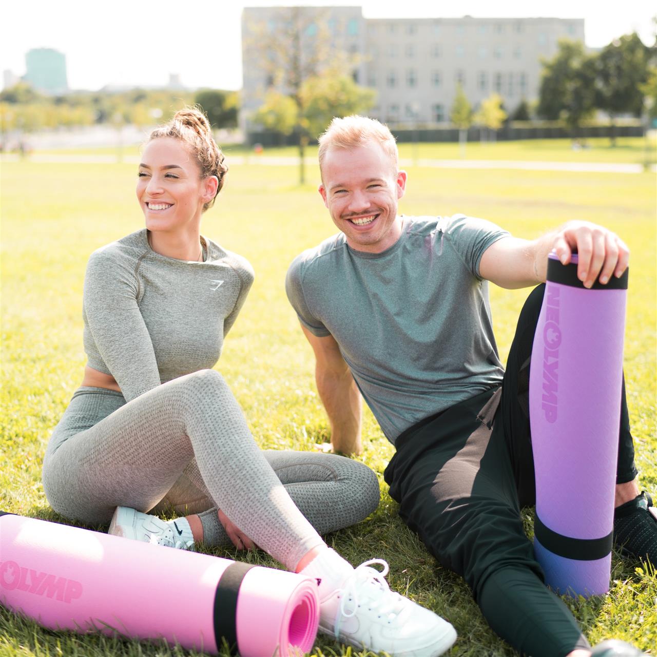 Zwei Personen sitzen lächelnd auf einer Wiese im Freien. Die Frau trägt ein graues Sportoutfit und hält eine rosa Yogamatte, während der Mann in einem grauen T-Shirt und schwarzen Hosen eine lilafarbene Yogamatte von NEOLYMP hält. Im Hintergrund sind unscharf ein paar Bäume und ein Gebäude zu sehen.