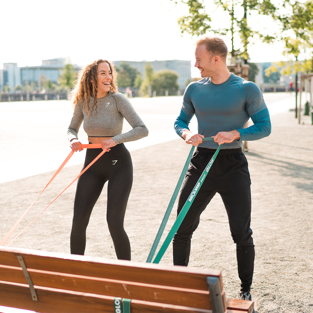 Zwei Personen trainieren im Freien mit Neolymp Fitnessbändern, die an einer Parkbank befestigt sind. Die Frau trägt ein graues Langarmshirt und schwarze Leggings, der Mann ein blaues Langarmshirt und schwarze Trainingshosen. Beide lachen und führen synchron Übungen mit den Fitnessbändern durch. Im Hintergrund sind Bäume und moderne Gebäude zu sehen, die Umgebung wirkt hell und sonnig.