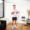 Ein Mann führt eine Übung mit einem Neolymp Fitnessband in einem modernen Wohnzimmer durch. Er trägt ein weißes T-Shirt, schwarze Shorts und weiße Sportschuhe. Im Hintergrund sind eine Treppe, eine schwarze Couch, ein Stuhl, ein kleines Regal und dekorative Pflanzen zu sehen.