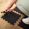 Eine Person setzt schwarze Puzzlematten mit roten Sprenkeln auf einem Holzboden zusammen. Die Matten bieten eine rutschfeste, schützende Oberfläche für Fitness- und Trainingsbereiche und sind einfach zu verlegen und anzupassen.